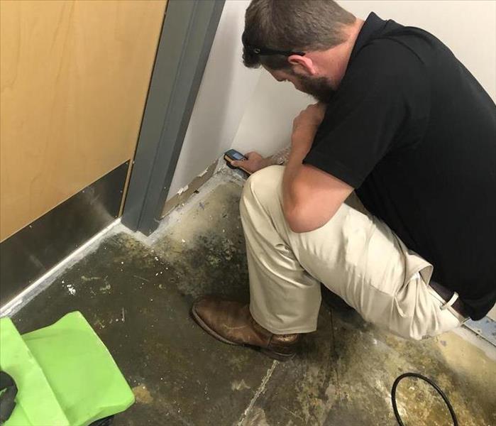 A male wearing a black polo and khaki pants, bent down taking moisture readings on a wall