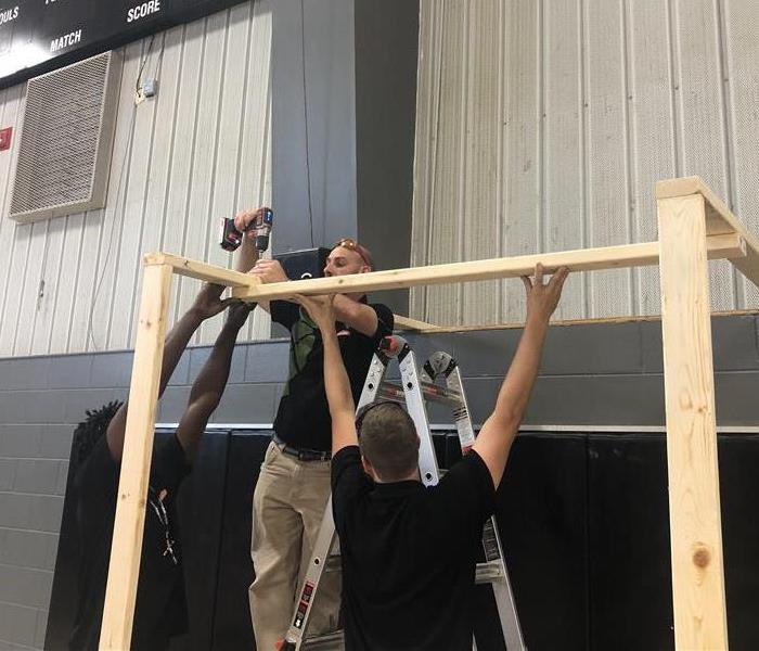 Three males in a gymnasium using wood to put together a small structure to make containment